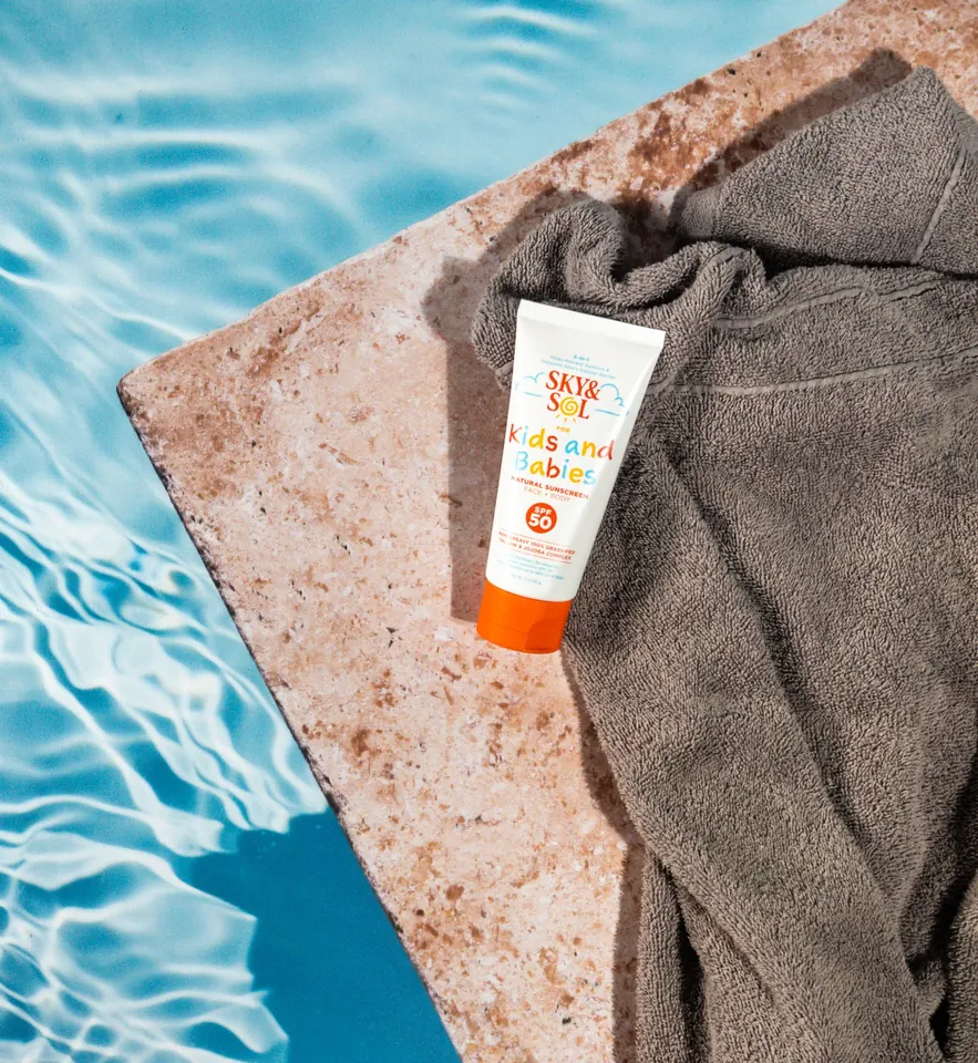 A tube of Sky and Sol sunscreen for kids and babies on a towel at the edge of a pool