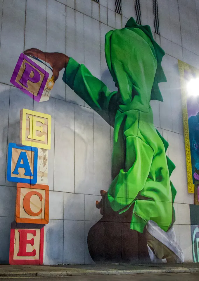 A street mural of a figure in a green hoodie stacking wooden blocks that spell peace
