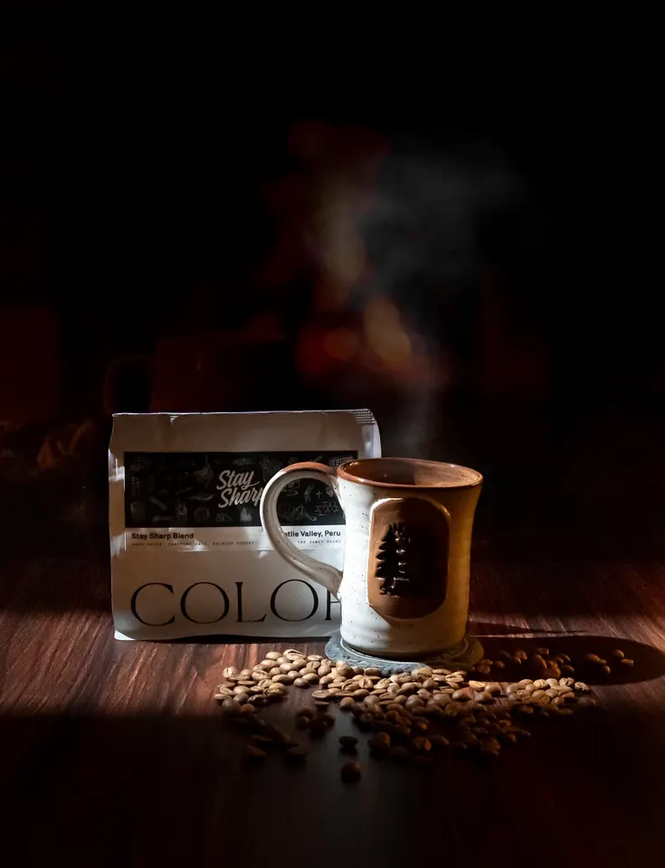 A steaming mug of coffee beside a bag of beans in low, warm light