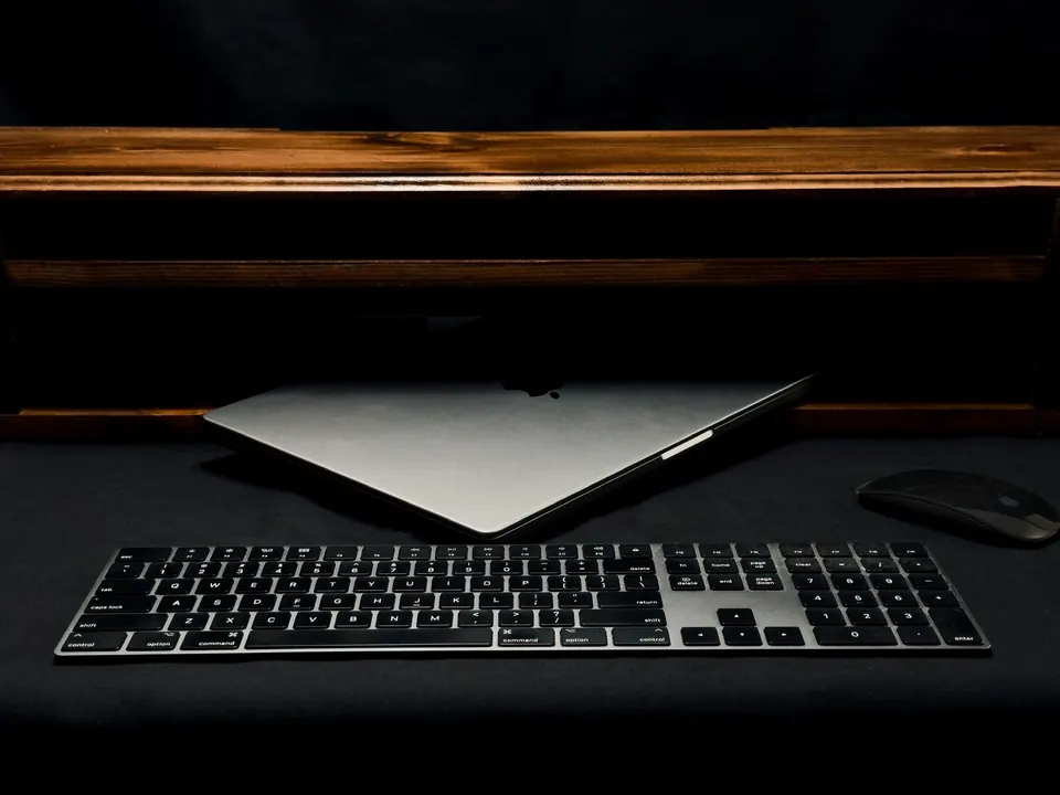 A laptop and keyboard on a dark desk, lit by a narrow strip of light