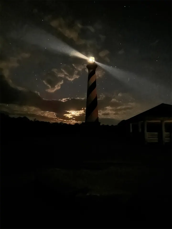 The Cape Hatteras lighthouse at night, its beams sweeping across a starry sky and moonlit clouds