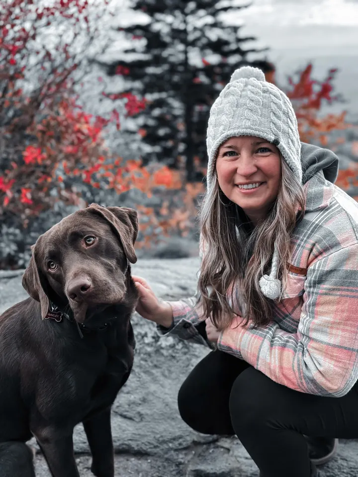 A smiling woman in a knit hat crouching beside a chocolate Labrador