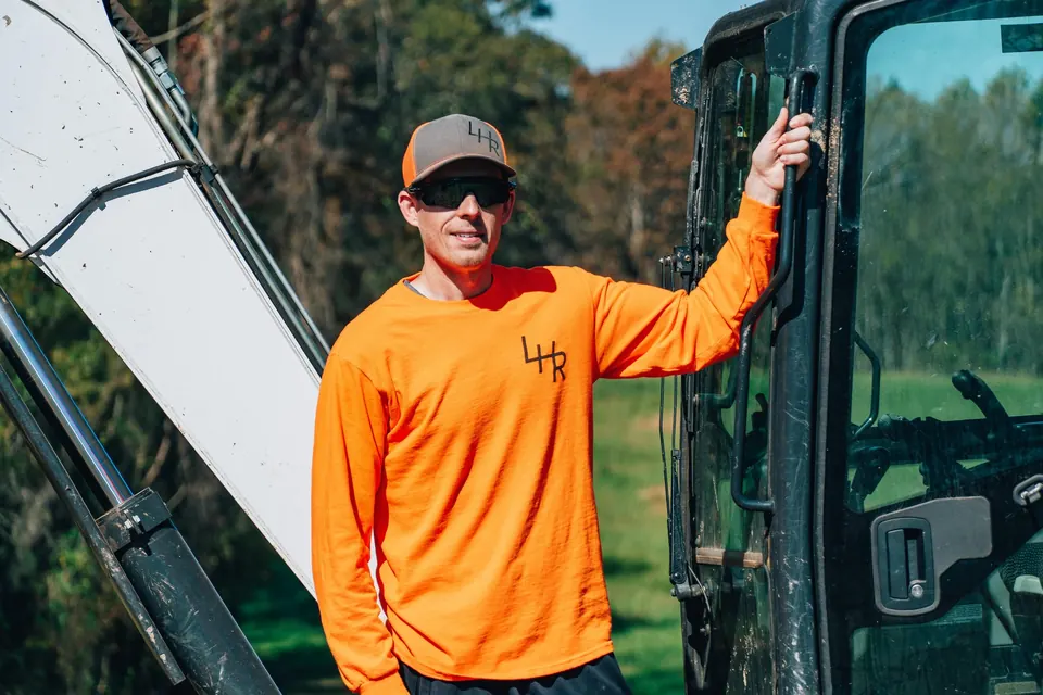 A man in an orange work shirt leaning out from the cab of an excavator